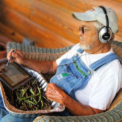 An old man with a large headset sits in a wicker chair using his ipad. On his lap is a large aluminum tray filled with asparagus