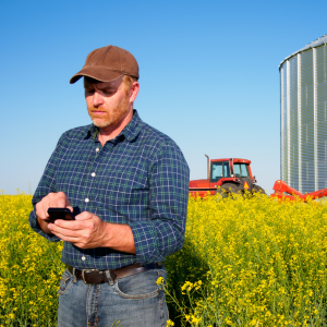 A farm worker checks his phone in a field, in the background a silo and a red tractor are visible. 